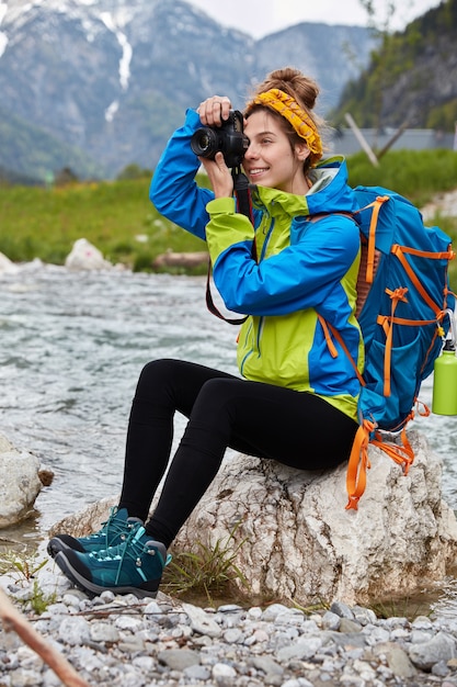 Pionowe ujęcie plenerowe wesołej samicy robi profesjonalne zdjęcia, siedzi na skałach w pobliżu górskiej rzeki