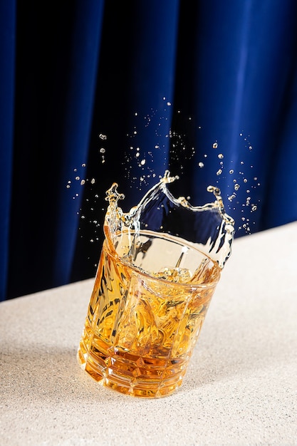 Bezpłatne zdjęcie pionowe ujęcie rozpryskującej się whisky w szklance z niebieską zasłoną w tle