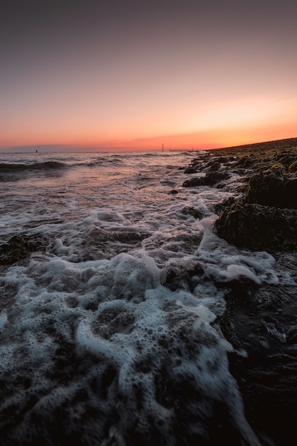 Pionowe ujęcie spienionych fal morza dochodzących do brzegu z niesamowitym zachodem słońca