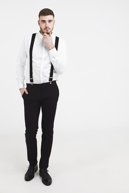 Pionowe ujęcie studyjne pełnej długości eleganckiego, przystojnego młodego brodatego modelu męskiego w czarnych spodniach, butach i białej koszuli z szelkami dotykającymi jego zarostu, pozujące do katalogu odzieży męskiej