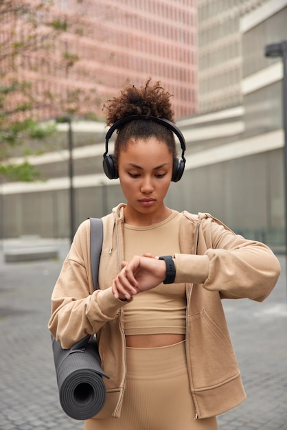 Pionowe ujęcie wysportowanej kobiety sprawdza wyniki fitness na smartwatchu monitoruje aktywność fizyczną ubraną w beżową odzież sportową niesie zwinięty karemat słucha muzyki przez słuchawki ćwiczenia na świeżym powietrzu