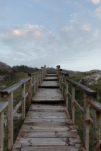 Pionowe zdjęcie drewnianych schodów w polu otoczonym zielenią i wzgórzami pod zachmurzonym niebem