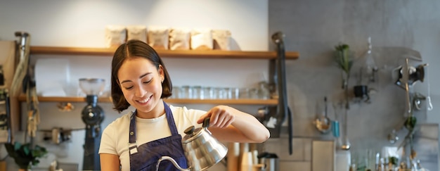 Pionowy strzał uśmiechniętego azjatyckiego barmana baristę w niebieskim fartuchu wylewającego wodę z małym czajnikiem