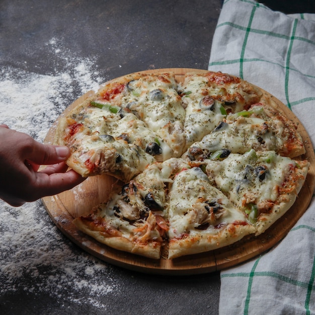 Pizza z boku na drewnianym stojaku z obrusem, ręka weź kawałek pizzy