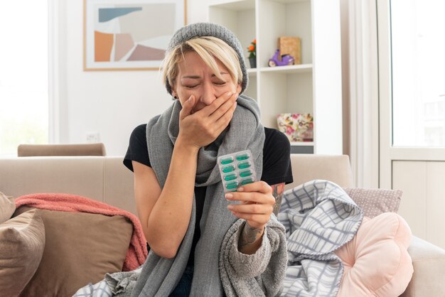 płacząca młoda chora słowiańska kobieta z szalikiem na szyi w czapce zimowej kładąca rękę na ustach i trzymająca blistry z lekarstwami siedząca na kanapie w salonie