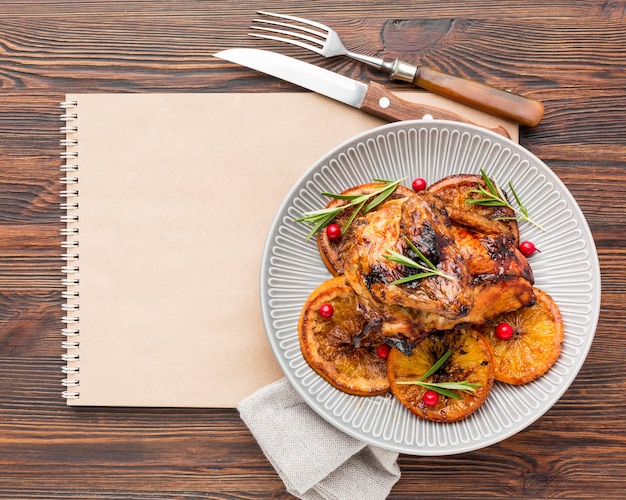 Płasko ułożony pieczony kurczak i plastry pomarańczy na talerzu ze sztućcami i pustym notatnikiem