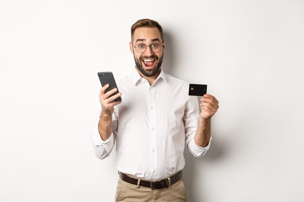 Płatności biznesowe i internetowe. Podekscytowany mężczyzna płacący telefonem komórkowym i kartą kredytową, uśmiechnięty zdumiony, stojący