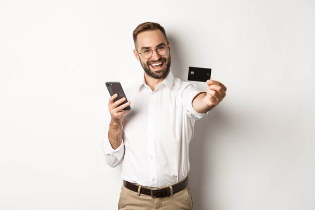 Płatności biznesowe i internetowe. Podekscytowany mężczyzna pokazuje swoją kartę kredytową, trzymając smartfon, stoi zadowolony