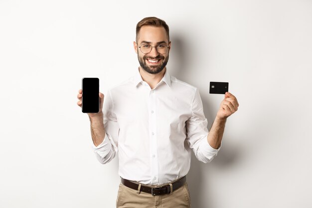 Płatności biznesowe i internetowe. Uśmiechnięty przystojny mężczyzna pokazuje ekran telefonu komórkowego i kartę kredytową, stojąc