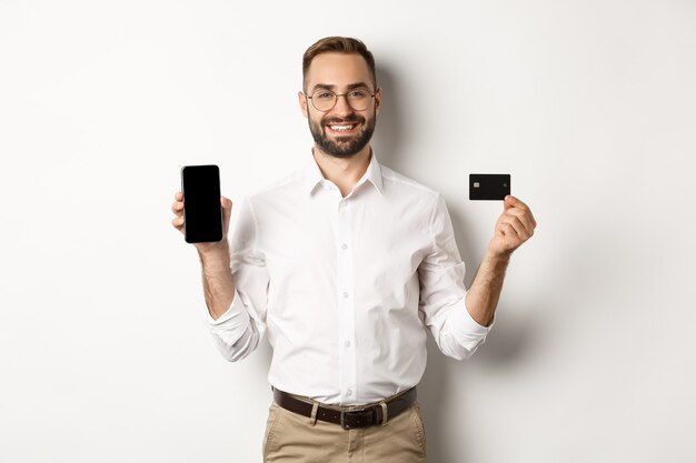 Płatności biznesowe i internetowe. Uśmiechnięty przystojny mężczyzna pokazuje ekran telefonu komórkowego i kartę kredytową, stojąc