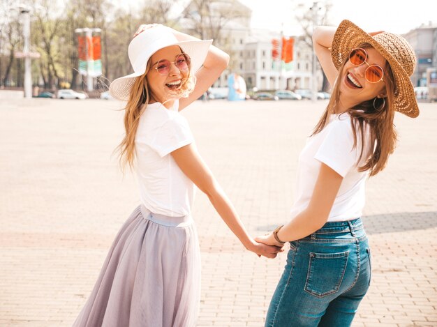 Plecy dwóch młodych pięknych blond uśmiechniętych hipster dziewcząt w modne letnie ubrania i kapelusz biały t-shirt. . Para trzymając się za ręce