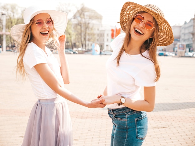 Plecy dwóch młodych pięknych blond uśmiechniętych hipster dziewcząt w modne letnie ubrania i kapelusz biały t-shirt. . Para trzymając się za ręce