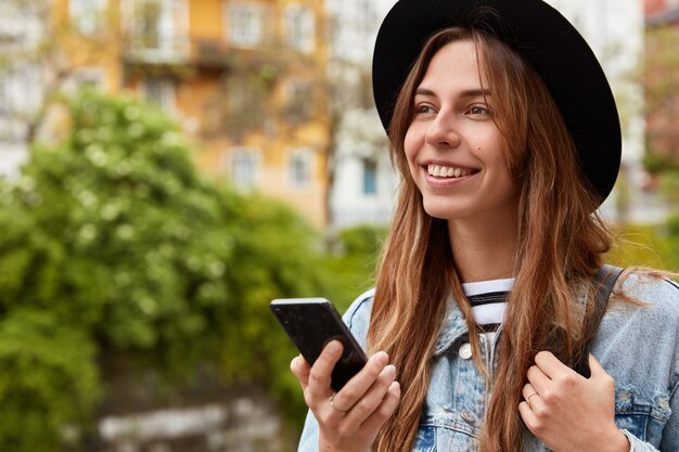 Plenerowe ujęcie rozmarzonej europejskiej kobiety w sieciach społecznościowych, spacery po mieście nad zieloną plantacją, z pozytywnym uśmiechem na twarzy