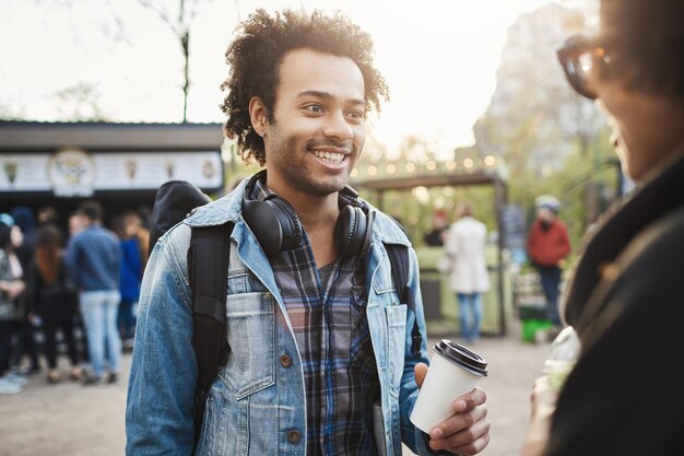 Plenerowy portret stylowego optymistycznego afrykańskiego mężczyzny z włosiem i fryzurą afro, trzymający filiżankę kawy i szeroko uśmiechnięty podczas rozmowy z atrakcyjną kobietą w parku