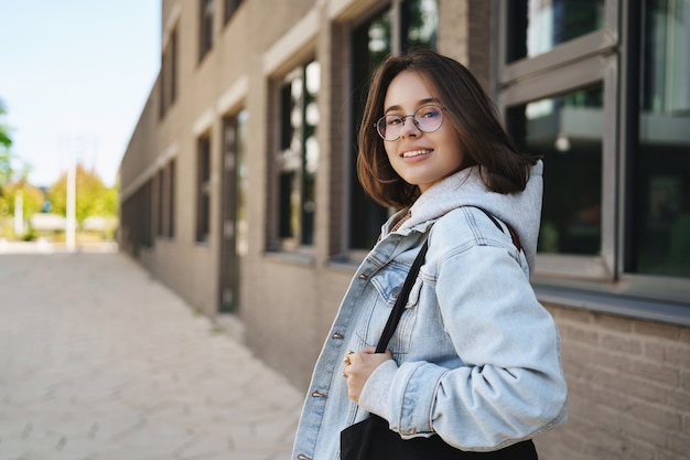 Plenerowy portret współczesnej młodej dziewczyny queer, studentki w okularach i dżinsowej kurtce, idącej do domu po zajęciach, odwracającej się z uśmiechem do kamery, czekającej na przyjaciela spacerującego po słonecznej ulicy.