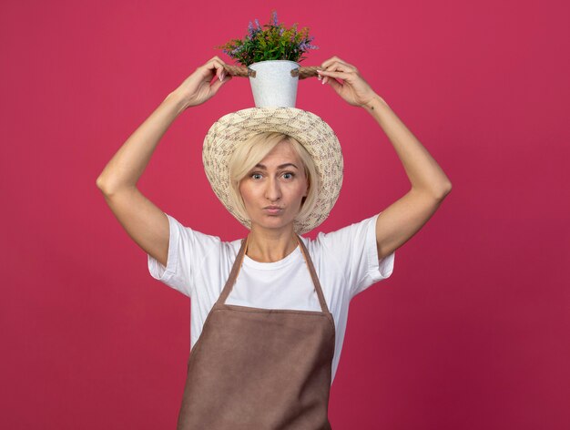 Bezpłatne zdjęcie pod wrażeniem blond ogrodniczka w średnim wieku w mundurze w kapeluszu trzymającym doniczkę na głowie odizolowaną na szkarłatnej ścianie