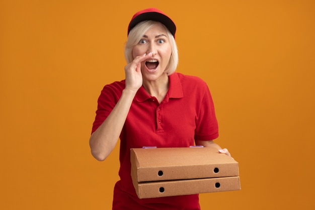 Bezpłatne zdjęcie pod wrażeniem blondyna w średnim wieku w czerwonym mundurze i czapce, trzymająca paczki z pizzą, trzymająca rękę przy ustach szepcząca