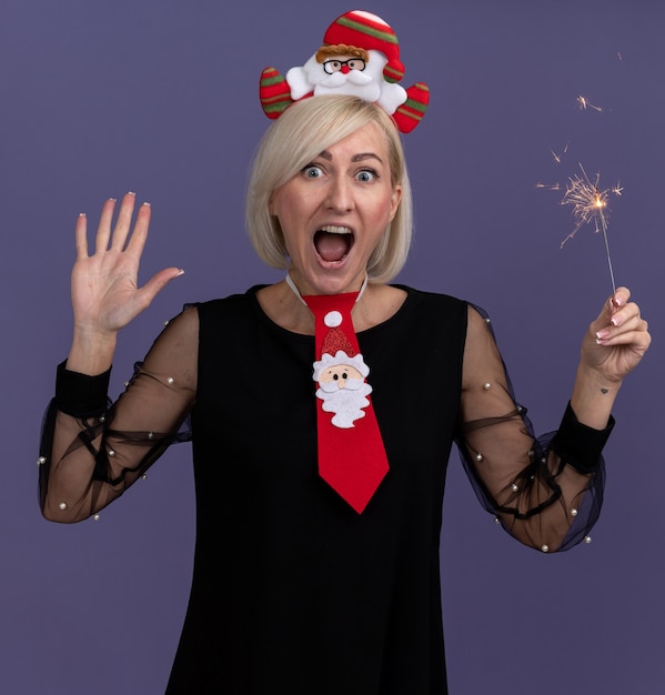 Bezpłatne zdjęcie pod wrażeniem blondynki w średnim wieku ubrana w opaskę świętego mikołaja i krawat trzyma świąteczny brylant patrząc na kamerę pokazującą pięć z ręką odizolowaną na fioletowym tle
