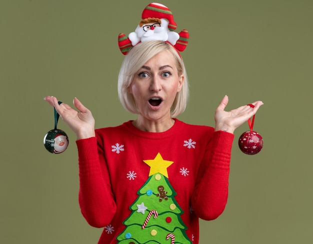 Pod wrażeniem blondynki w średnim wieku ubrana w opaskę świętego mikołaja i świąteczny sweter trzymający świąteczne bombki odizolowane na oliwkowej ścianie