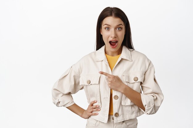 Pod wrażeniem brunetka w dżinsowej kurtce, zafascynowana otwartymi ustami, wskazująca palcem na sprzedaż po lewej stronie, logo lub kopia miejsca na bok