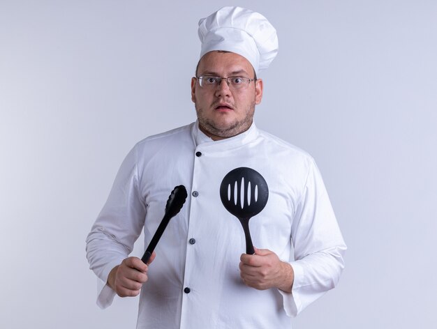 pod wrażeniem dorosłego kucharza w mundurze szefa kuchni i okularach, trzymających szczypce i łyżkę cedzakową, patrząc na przód na białej ścianie
