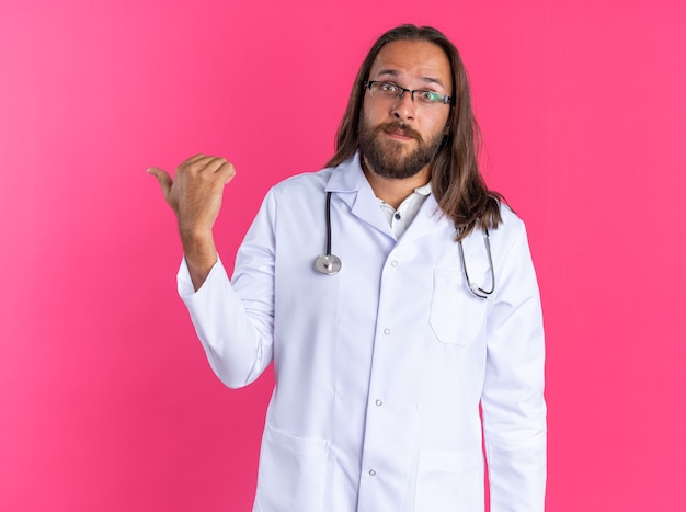 Pod wrażeniem dorosłego lekarza płci męskiej noszącego szatę medyczną i stetoskop w okularach, patrząc na kamerę wskazującą na bok odizolowaną na różowej ścianie