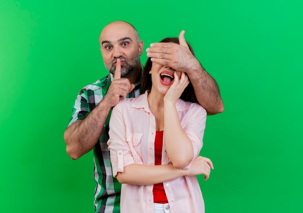 Bezpłatne zdjęcie pod wrażeniem dorosłej pary mężczyzna zakrywający oczy kobiety ręką gestykulującą ciszę kobieta kładąca dłoń na twarzy odizolowana na zielonej ścianie z przestrzenią do kopiowania