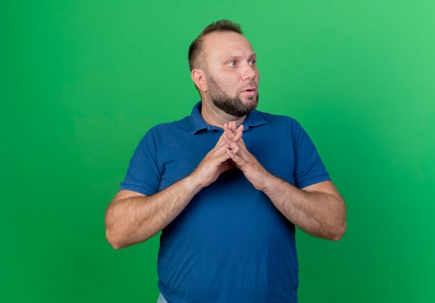 Pod wrażeniem dorosły mężczyzna słowiański patrząc na bok, trzymając ręce razem na białym tle na zielonej ścianie z miejsca na kopię