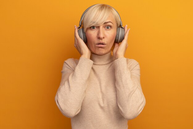 Pod wrażeniem ładna blondynka słowiańska kobieta na słuchawkach patrzy na aparat na pomarańczowo