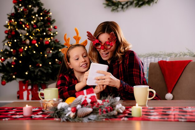 Pod wrażeniem matka w okularach renifera patrząc na notebooka z córką siedzącą przy stole, ciesząc się świątecznym w domu