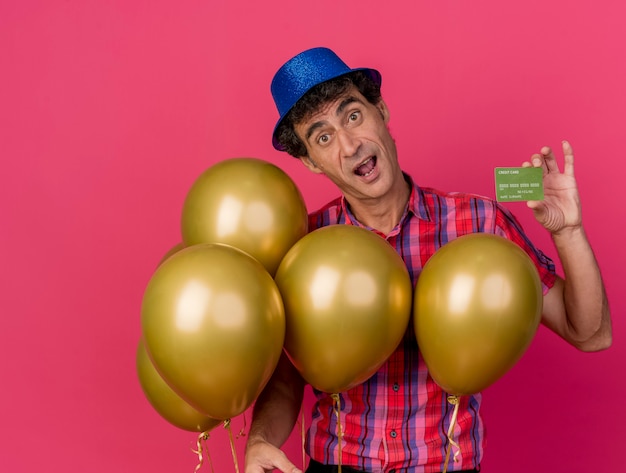 Bezpłatne zdjęcie pod wrażeniem mężczyzna w średnim wieku strona ubrana w kapelusz partii trzymając balony pokazując kartę kredytową patrząc z przodu na białym tle na szkarłatnej ścianie