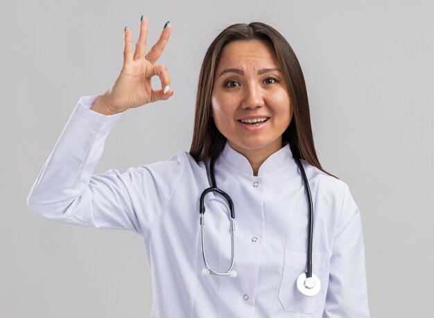 Pod wrażeniem młoda azjatycka lekarka nosząca medyczną szatę i stetoskop, patrząc na przód, robiąc znak ok na białej ścianie
