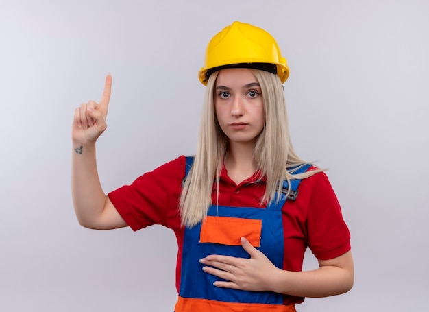 Bezpłatne zdjęcie pod wrażeniem młoda blondynka inżynier konstruktor dziewczyna w mundurze skierowana w górę ręką na brzuchu na odosobnionej białej przestrzeni