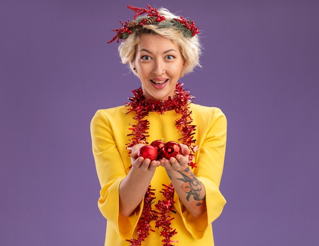 Bezpłatne zdjęcie pod wrażeniem młoda blondynka ubrana w świąteczny wieniec na głowę i świecącą girlandę na szyi, patrząc trzymając bombki świąteczne na fioletowej ścianie z miejscem na kopię