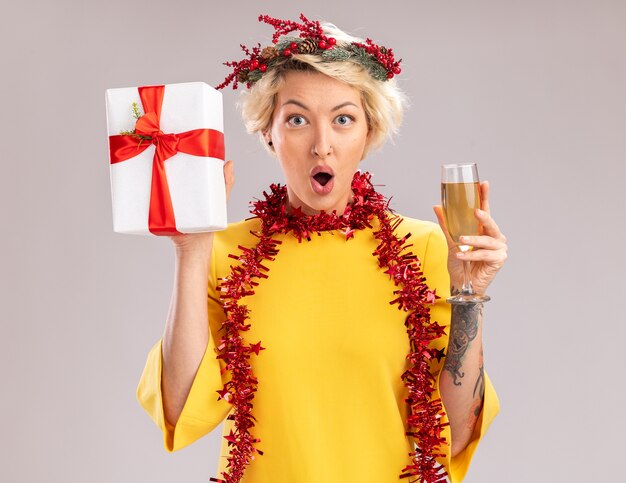 Pod wrażeniem młoda blondynka ubrana w świąteczny wieniec na głowę i świecącą girlandę na szyi trzyma kieliszek szampana i pakiet prezentów patrząc na aparat na białym tle