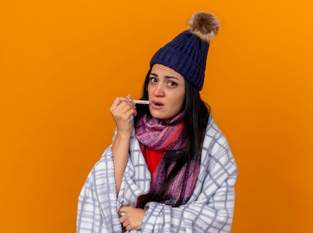 Bezpłatne zdjęcie pod wrażeniem młoda chora kobieta w czapce zimowej i szaliku owinięta w kratę wkładająca termometr do ust patrząc z przodu odizolowana na pomarańczowej ścianie