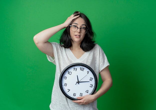Pod wrażeniem młoda dziewczyna całkiem kaukaski w okularach, trzymając zegar i kładąc rękę na głowie na białym tle na zielonym tle z miejsca na kopię