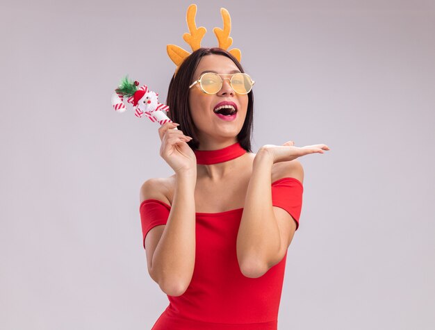 Pod wrażeniem młoda dziewczyna nosi opaskę z poroża renifera i okulary, trzymając ozdobę świątecznej trzciny cukrowej, patrząc na kamery pokazując pustą rękę na białym tle