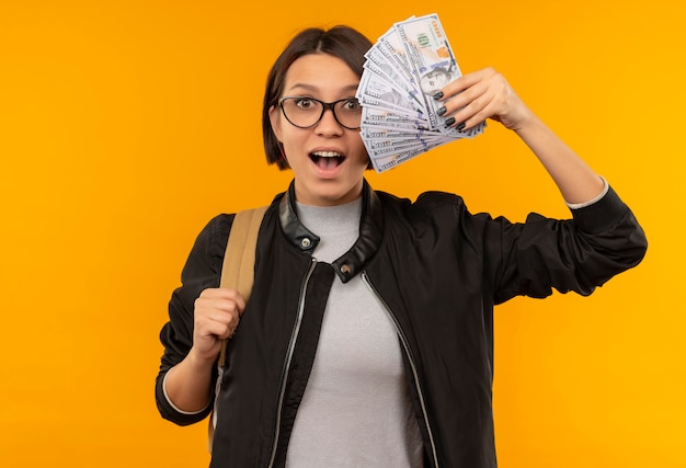 Pod wrażeniem młoda dziewczyna studenta w okularach iz powrotem worek trzymając pieniądze na białym tle na pomarańczowo