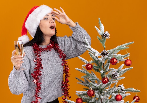 pod wrażeniem młoda kaukaska dziewczyna nosząca świąteczny kapelusz i blichtrową girlandę na szyi stojącą w pobliżu udekorowanej choinki trzymającej kieliszek szampana patrzący z boku w dal wyizolowaną na pomarańczowej ścianie