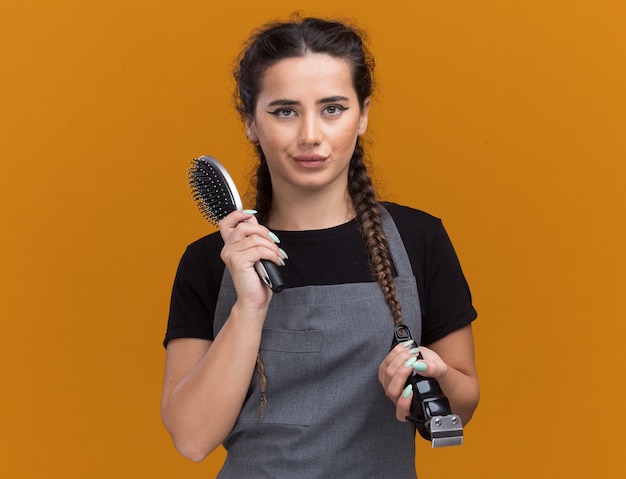 Pod wrażeniem młoda kobieta fryzjerka w mundurze trzymając grzebień z maszynką do strzyżenia włosów na białym tle na pomarańczowej ścianie