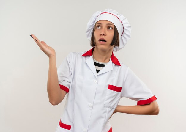 Pod wrażeniem młoda kobieta kucharz w mundurze szefa kuchni, patrząc na bok, pokazując pustą rękę i kładąc rękę na talii na białym tle