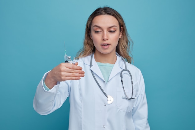 Pod wrażeniem młoda kobieta lekarz ubrana w szlafrok medyczny i stetoskop wokół szyi, trzymając i patrząc na ampułkę strzykawki i butelkę ze szczepionką