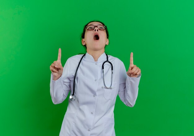 Pod wrażeniem młoda kobieta lekarz ubrany w medyczną szatę i stetoskop i okulary patrząc i skierowaną w górę na białym tle na zielonej ścianie z miejsca na kopię