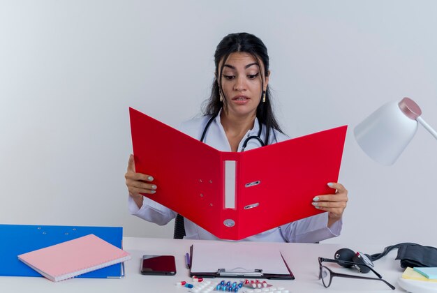 Pod wrażeniem młoda kobieta lekarz ubrany w medyczną szatę i stetoskop siedzi przy biurku z narzędzi medycznych, trzymając i patrząc na folder odizolowane