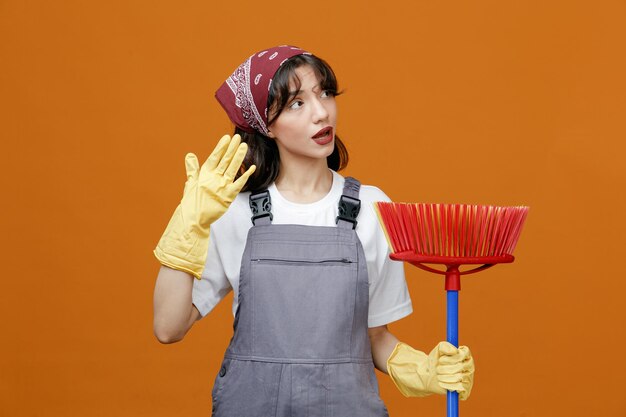 Pod wrażeniem młoda kobieta sprzątaczka w jednolitych gumowych rękawiczkach i chustce, trzymając rękę w powietrzu, trzymając mopa ściągaczki, patrząc na bok na białym tle na pomarańczowym tle