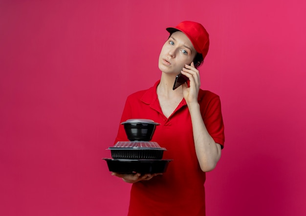 Pod wrażeniem młoda ładna dziewczyna dostawy ubrana w czerwony mundur i czapkę, rozmawiająca przez telefon i trzymająca pojemniki z jedzeniem, patrząc na kamerę odizolowaną na szkarłatnym tle z przestrzenią do kopiowania