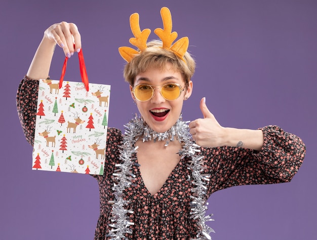 Bezpłatne zdjęcie pod wrażeniem młoda ładna dziewczyna ubrana w opaskę z poroża renifera i świecącą girlandę na szyi w okularach trzyma torbę z prezentami świątecznymi pokazując kciuk w górę na fioletowej ścianie