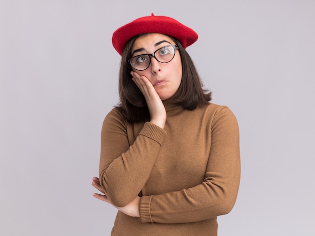 Pod wrażeniem młoda ładna kaukaska dziewczyna w beretowym kapeluszu i w okularach optycznych kładzie rękę na twarzy na białym tle na białej ścianie z kopią przestrzeni