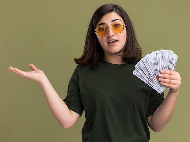 Pod wrażeniem młoda ładna kaukaska dziewczyna w okularach przeciwsłonecznych trzyma pieniądze i trzyma rękę otwartą na oliwkowej zieleni